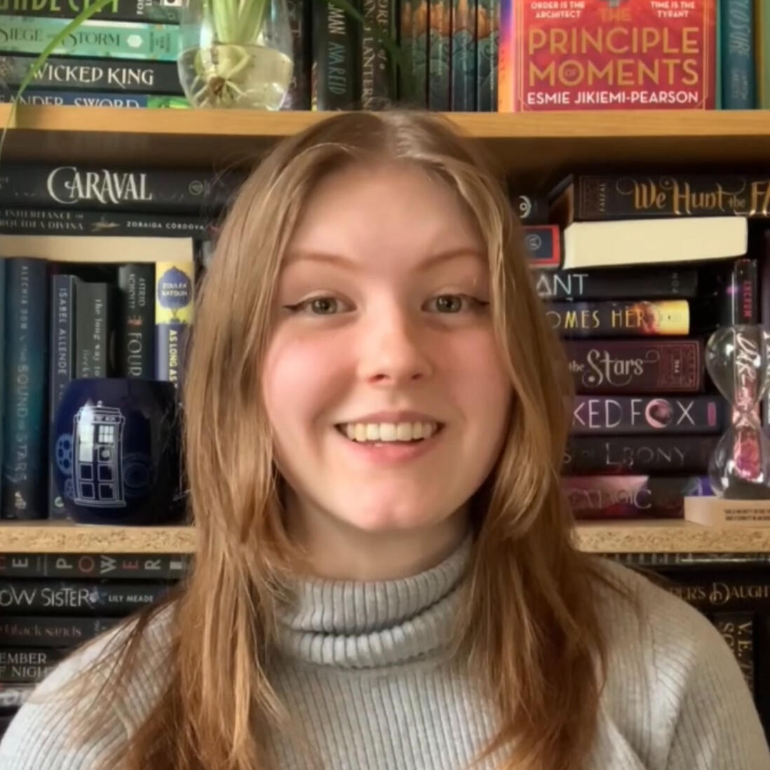 Ellie, a white woman with long reddish-blond hair wearing a gray turtleneck sweater, smiles in front of a colorful bookshelf.