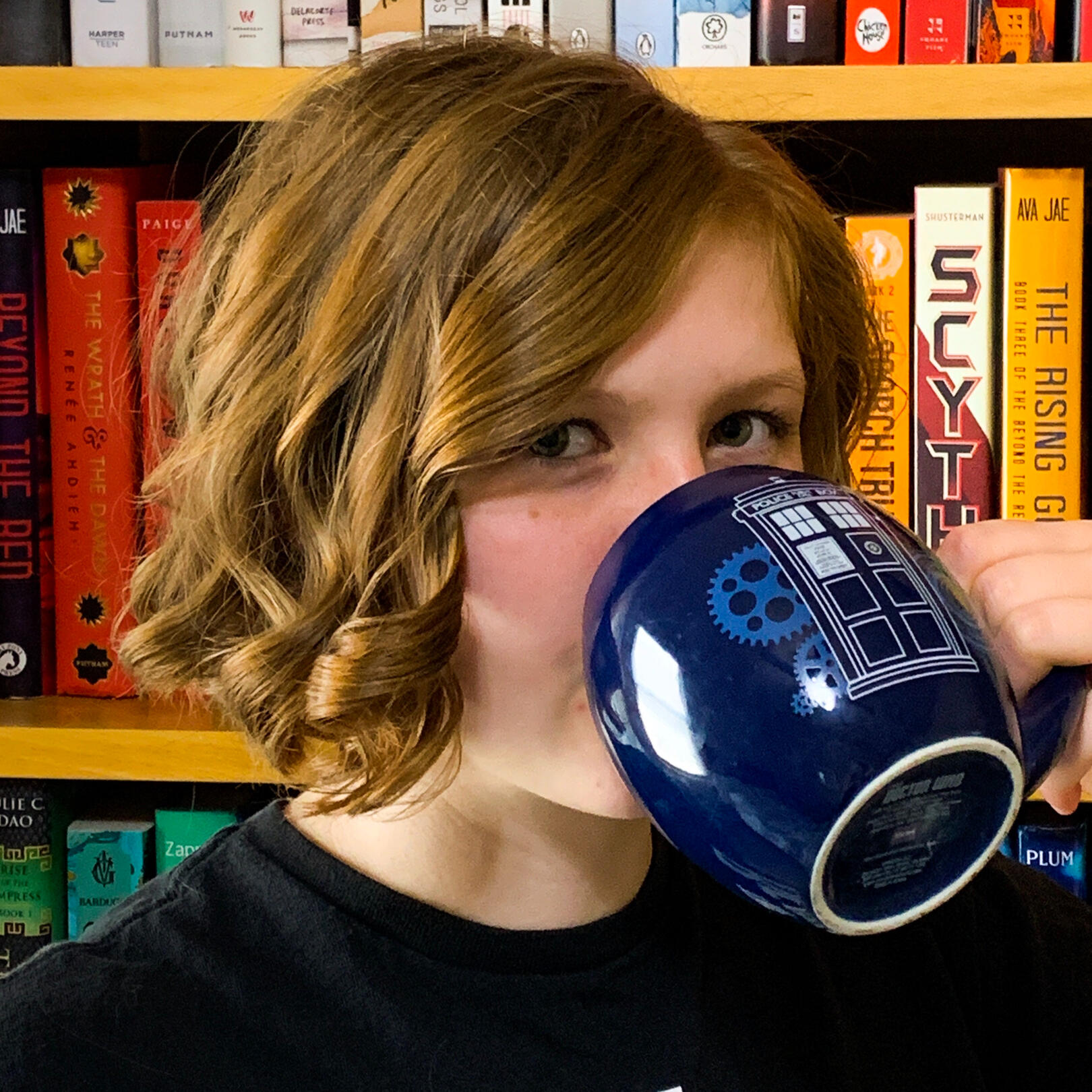 A white woman with short curly reddish-blond hair drinking out of a TARDIS mug with a colorful bookshelf behind her.